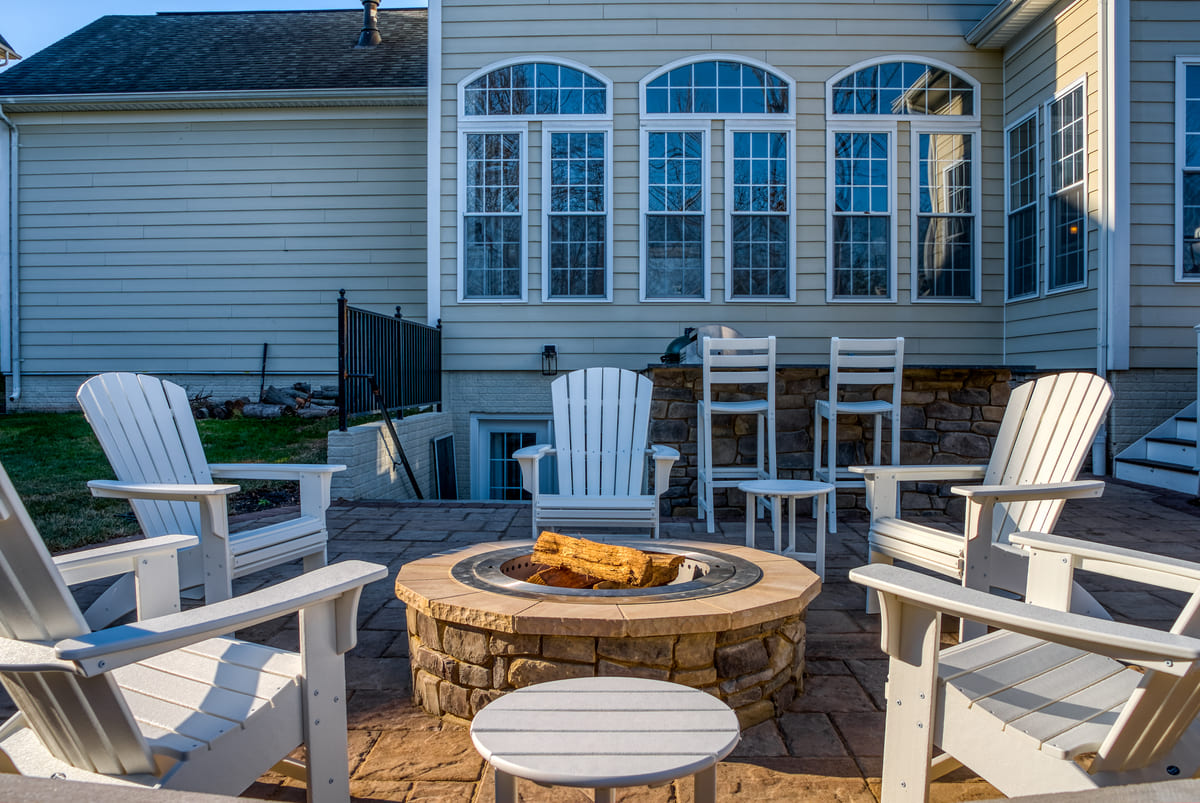 Adirondack chairs surrounding stone fire pit on patio with outdoor kitchen in back by Deckscapes of Virginia