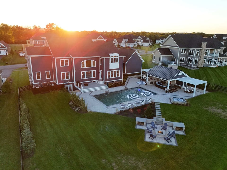 Sun setting over drone view of backyard remodel featuring patio, pool, fireput, fireplace, spa, pergola, outdoor kitchen, and more
