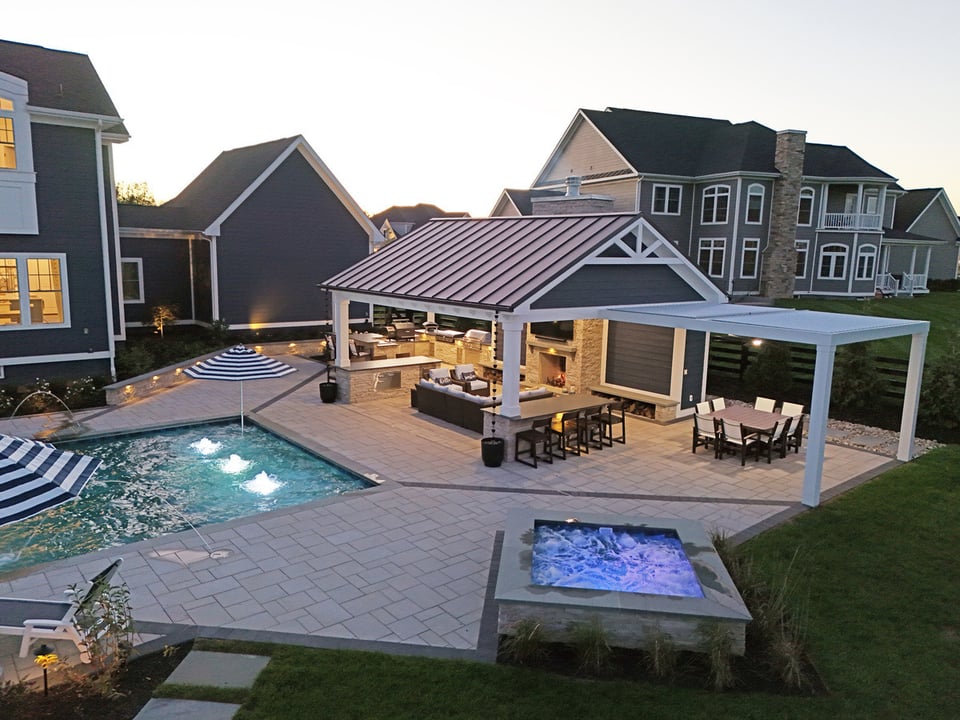 Backyard living space lit up at dusk with outdoor kitchen, covered seating, pool, and firepit