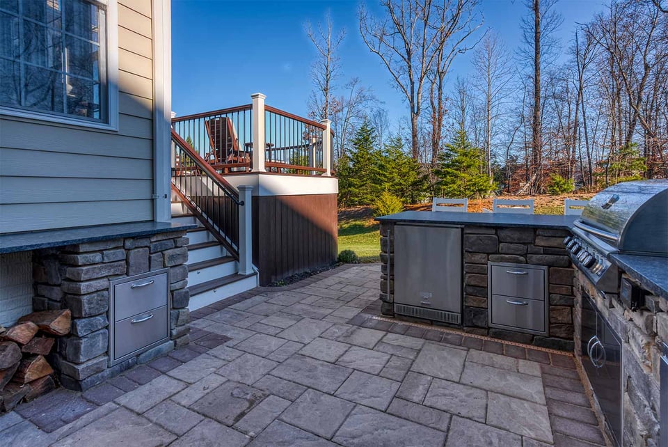 Outdoor kitchen with built-in grill, stone cabinetry, and deck stairs leading to backyard patio by Deckscapes of Virginia
