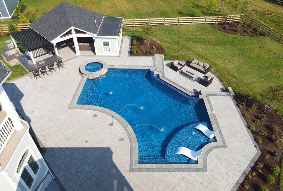 Modern backyard pool with fountains, lounge shelf, and pavilion with bar and firepit area by Deckscapes of Virginia