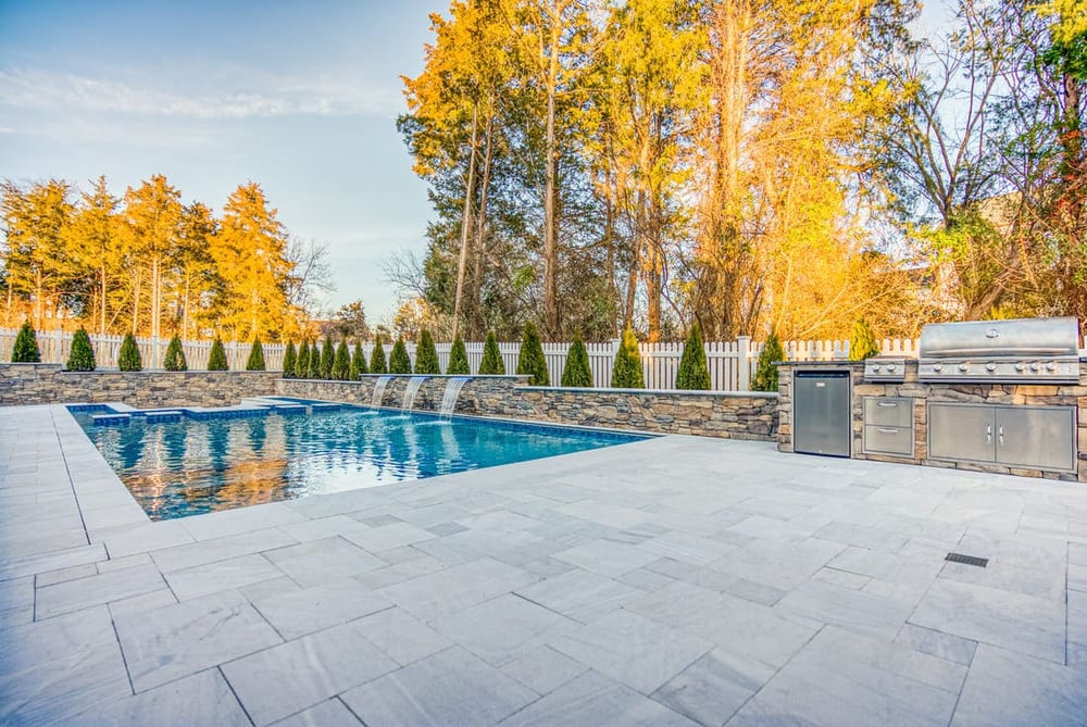 Marble pavers surrounding modern gunite pool with stacked stone outdoor kitchen by Deckscapes of VA
