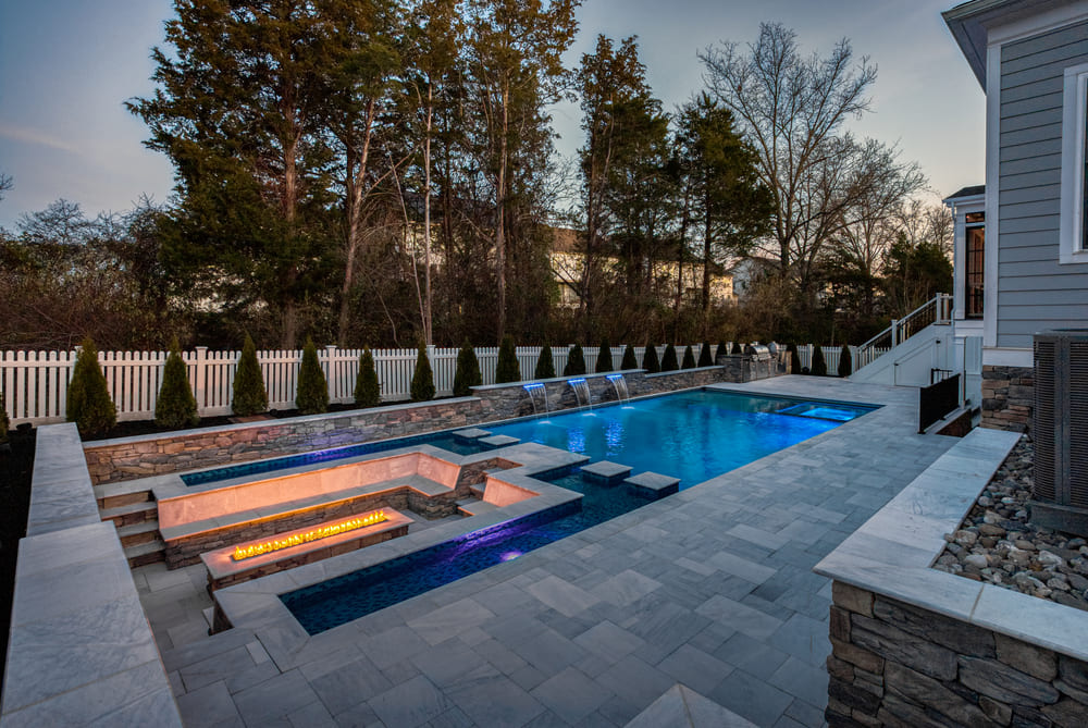 Northern VA backyard with gunite pool and fire pit at dusk with custom lighting installation by Deckscapes of VA