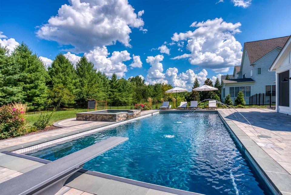 Backyard lap pool with diving board, fountains, and lounge chairs under umbrellas by Deckscapes of Virginia