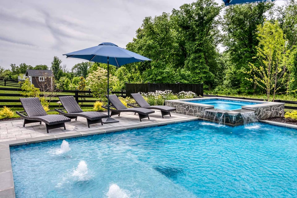 Backyard pool with stone spa, waterfalls, lounge chairs, and blue umbrella in lush garden setting by Deckscapes of Virginia