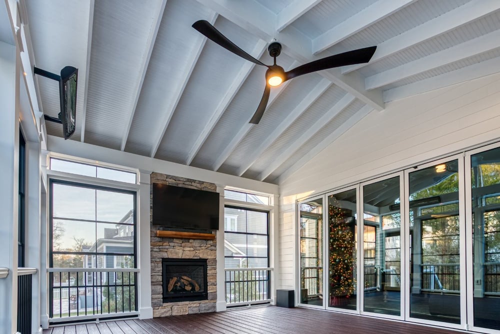 Screened-in porch with floating tv and stone fireplace below ceiling fan and Bromic heater