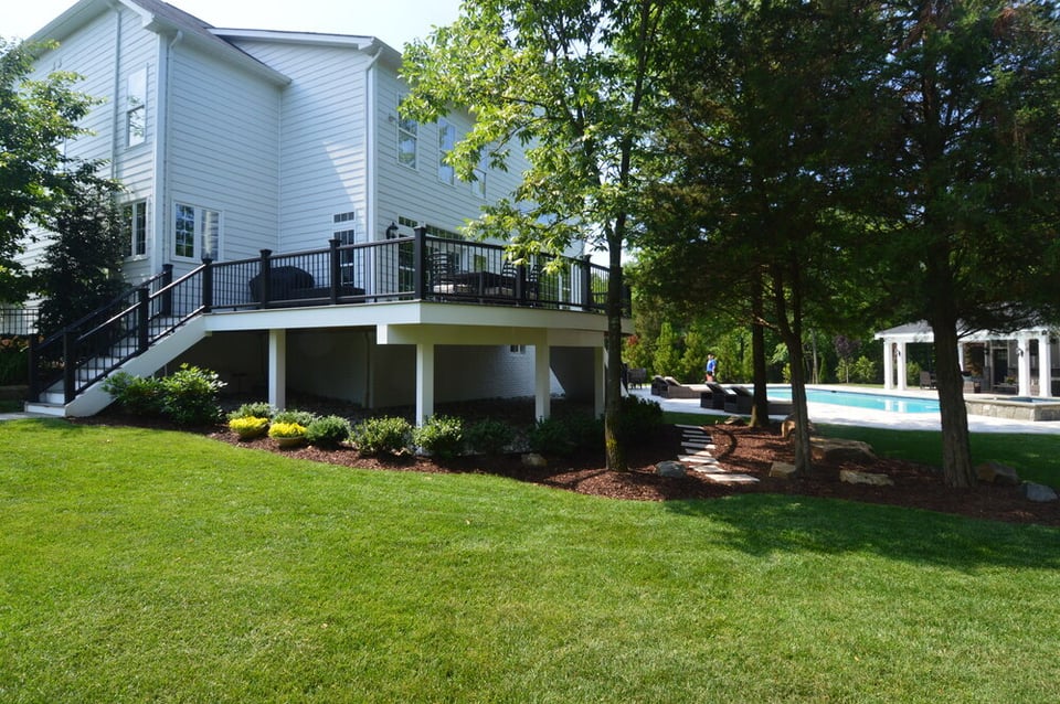 Wide shot of a second-story deck with two staircases and custom landscaping below pool and pool house beyond by Deckscapes of Virginia