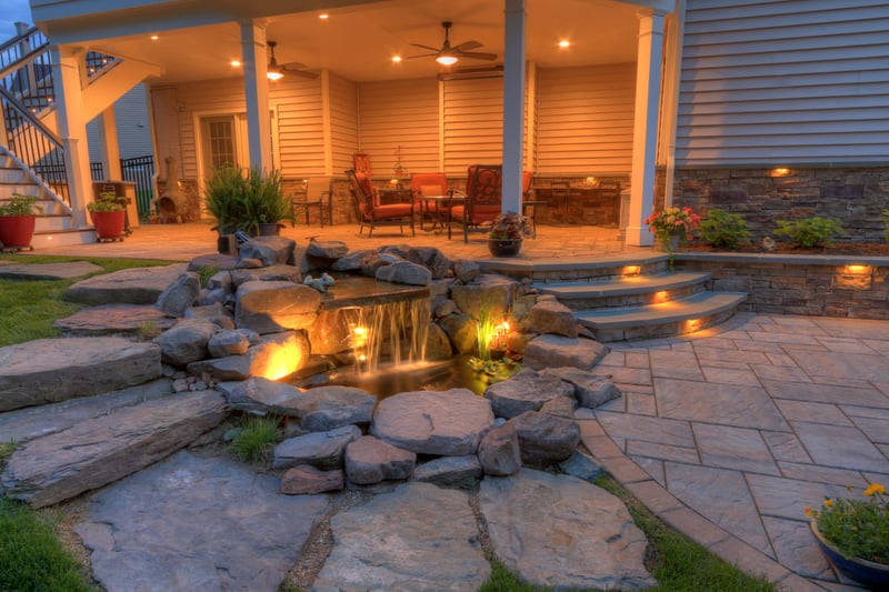 Waterfall feature next to stone pathway leading to patio at dusk with custom lighting installation by Deckscapes of Virginia