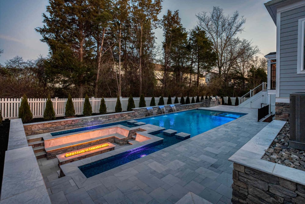 night time back yard with pool lights on and fire place surrounded by pool (1)